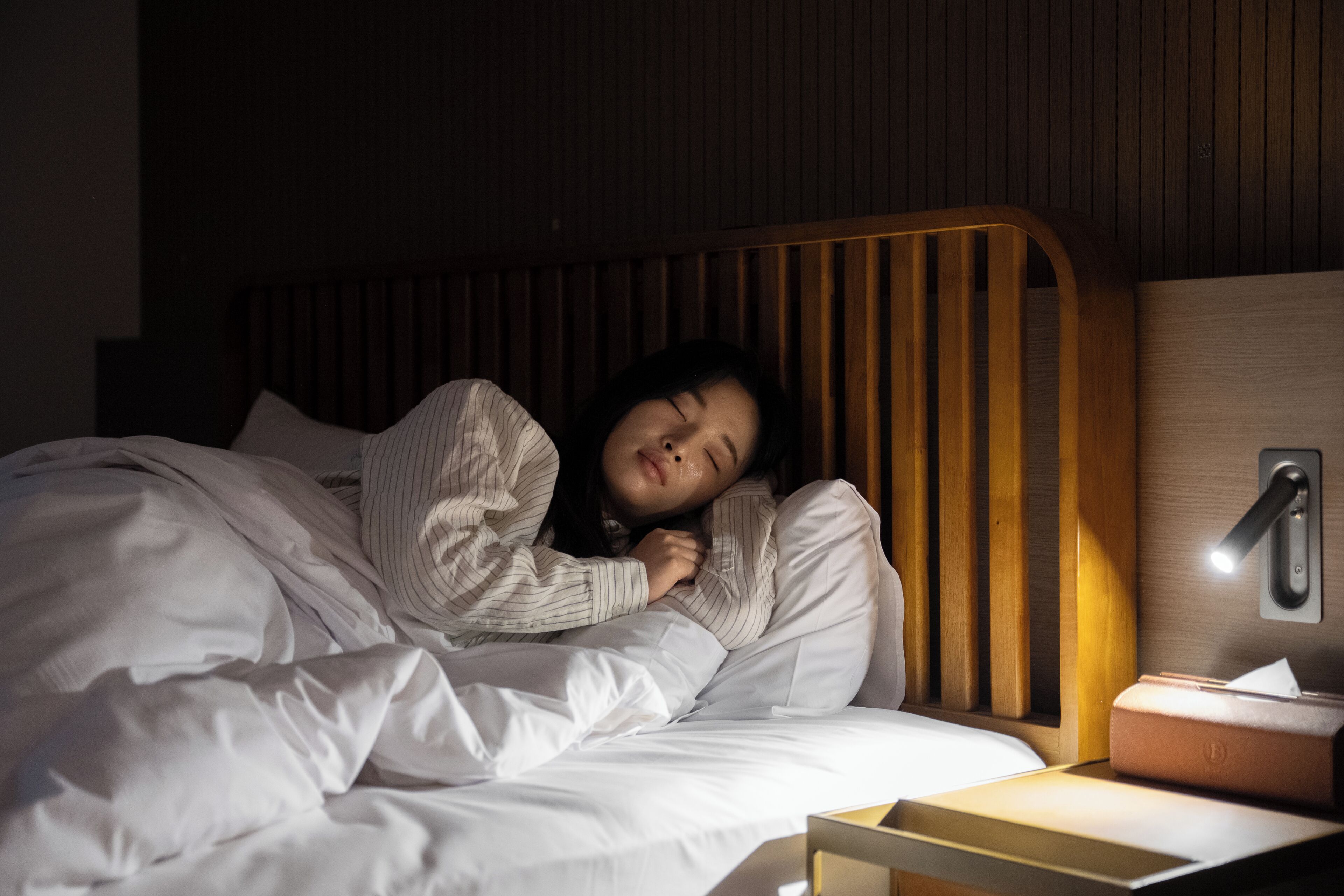 Une personne est allongée dans un lit doté d’une tête de lit en bois, d’une table de chevet avec une lampe et d’une boîte de mouchoirs.