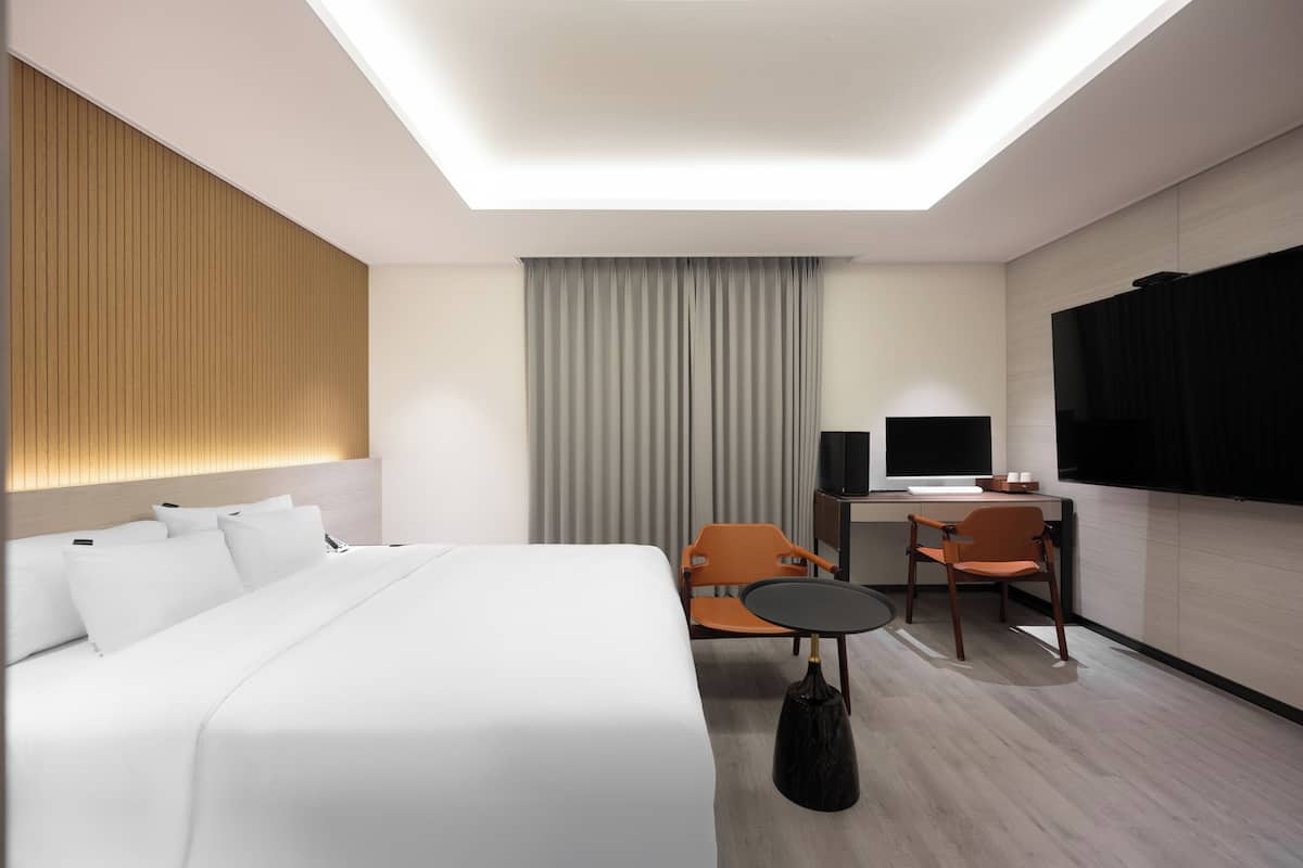A modern hotel room with a large bed, a desk with a computer, two chairs, a TV, and a ceiling light.