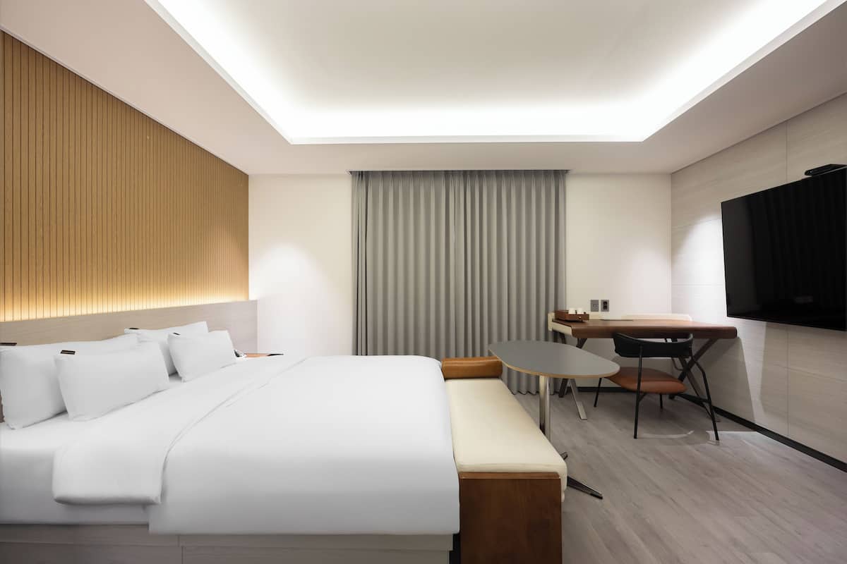 A modern hotel room with a bed, bedside table, chair, desk, and a flat-screen TV.