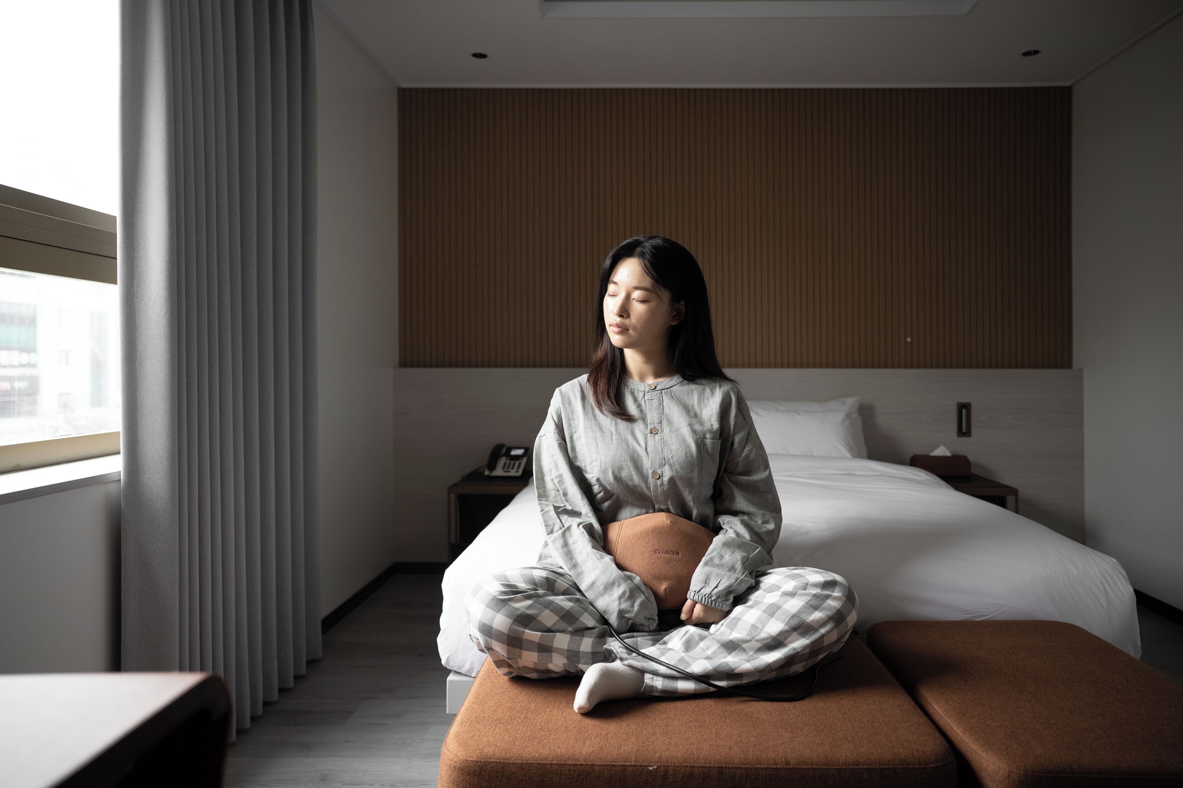 Une personne est assise sur un lit dans une chambre d’hôtel. On peut voir un mur avec des panneaux en bois, une table de chevet et une fenêtre avec des rideaux.