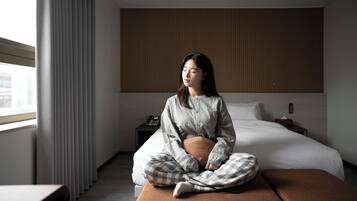 A person sitting on a bed in a hotel room with a wooden panel wall, a bedside table, and a window with curtains.