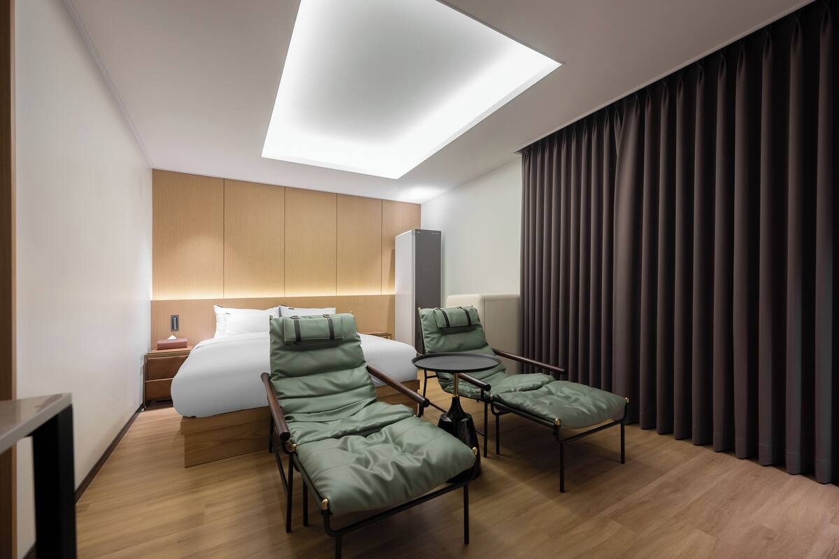 A modern hotel room with a skylight, a bed, two green lounge chairs, a small table, and a large window with curtains.