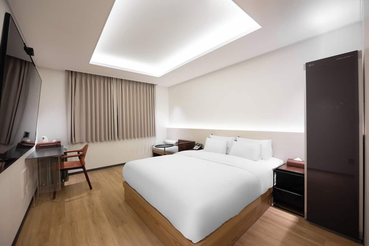 A modern hotel room with a large bed, a desk, a chair, a small table, a nightstand, and a wardrobe.