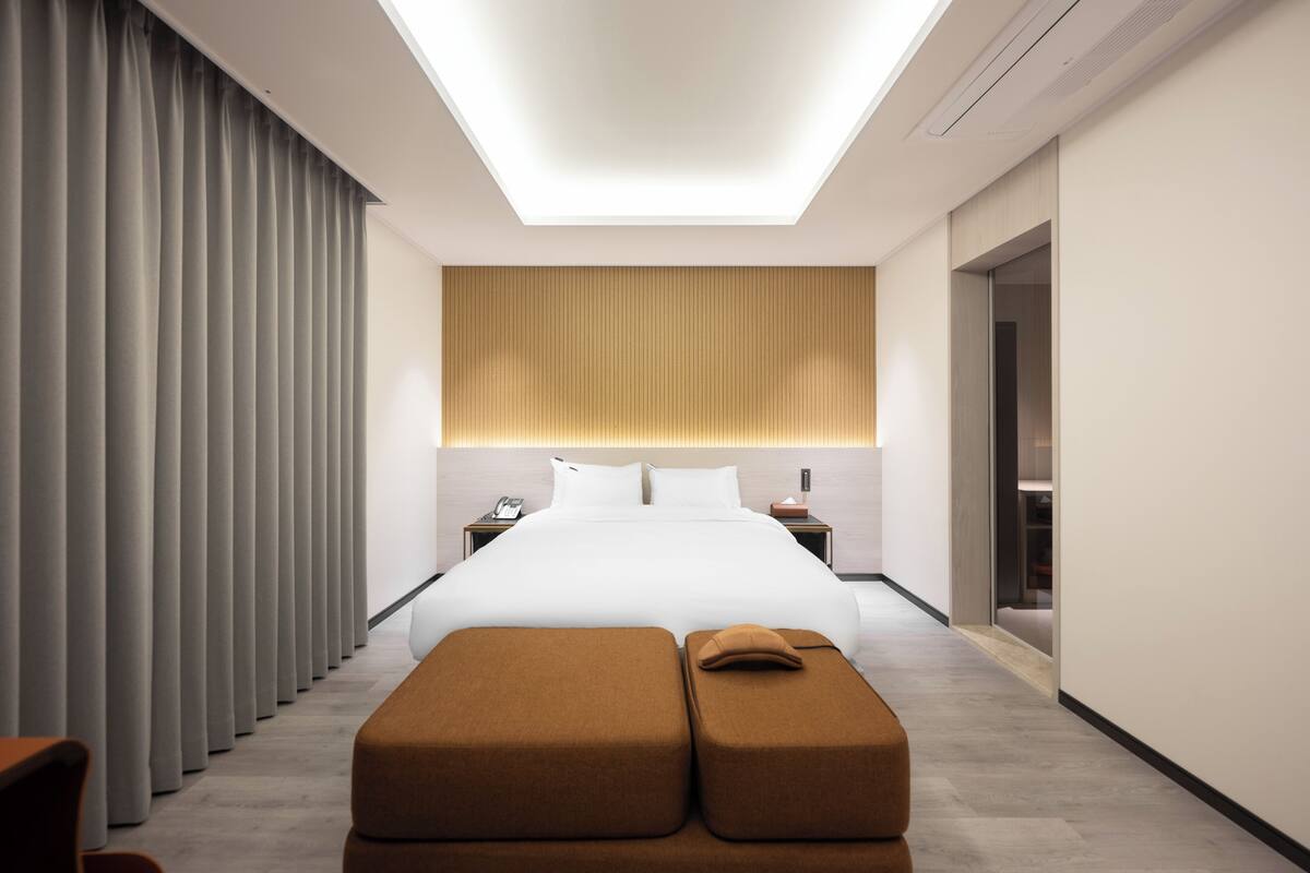 A modern hotel room with a large bed, a bedside table, a nightstand, and a window with curtains.