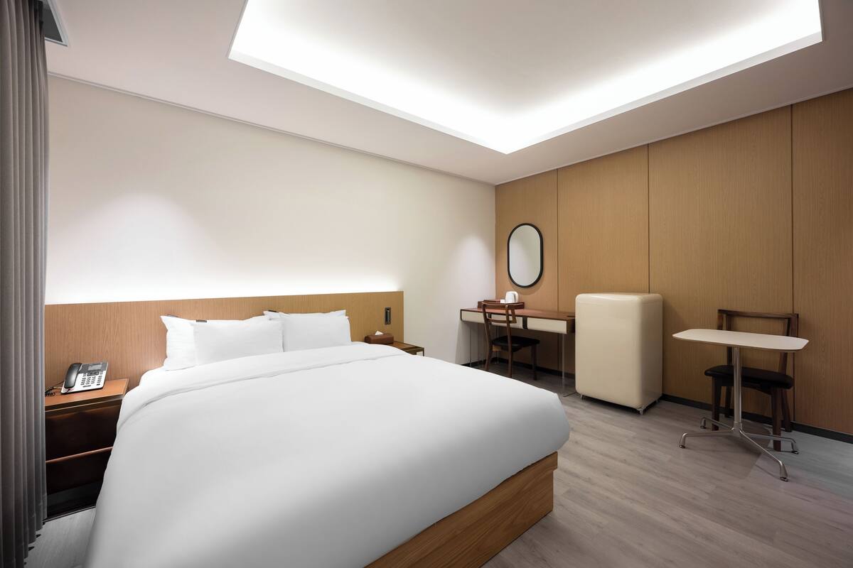 A hotel room with a bed, a desk, a chair, a small table, and a telephone on the nightstand.