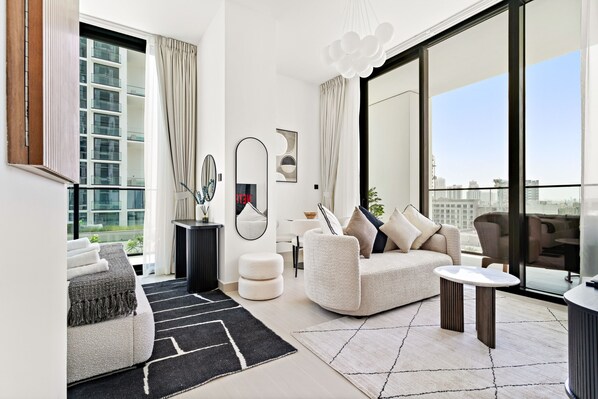 A modern living room with a sofa, ottoman, and coffee table, large windows with curtains, and a city view. - Spacious Modern Studio in JVC (Dubai)