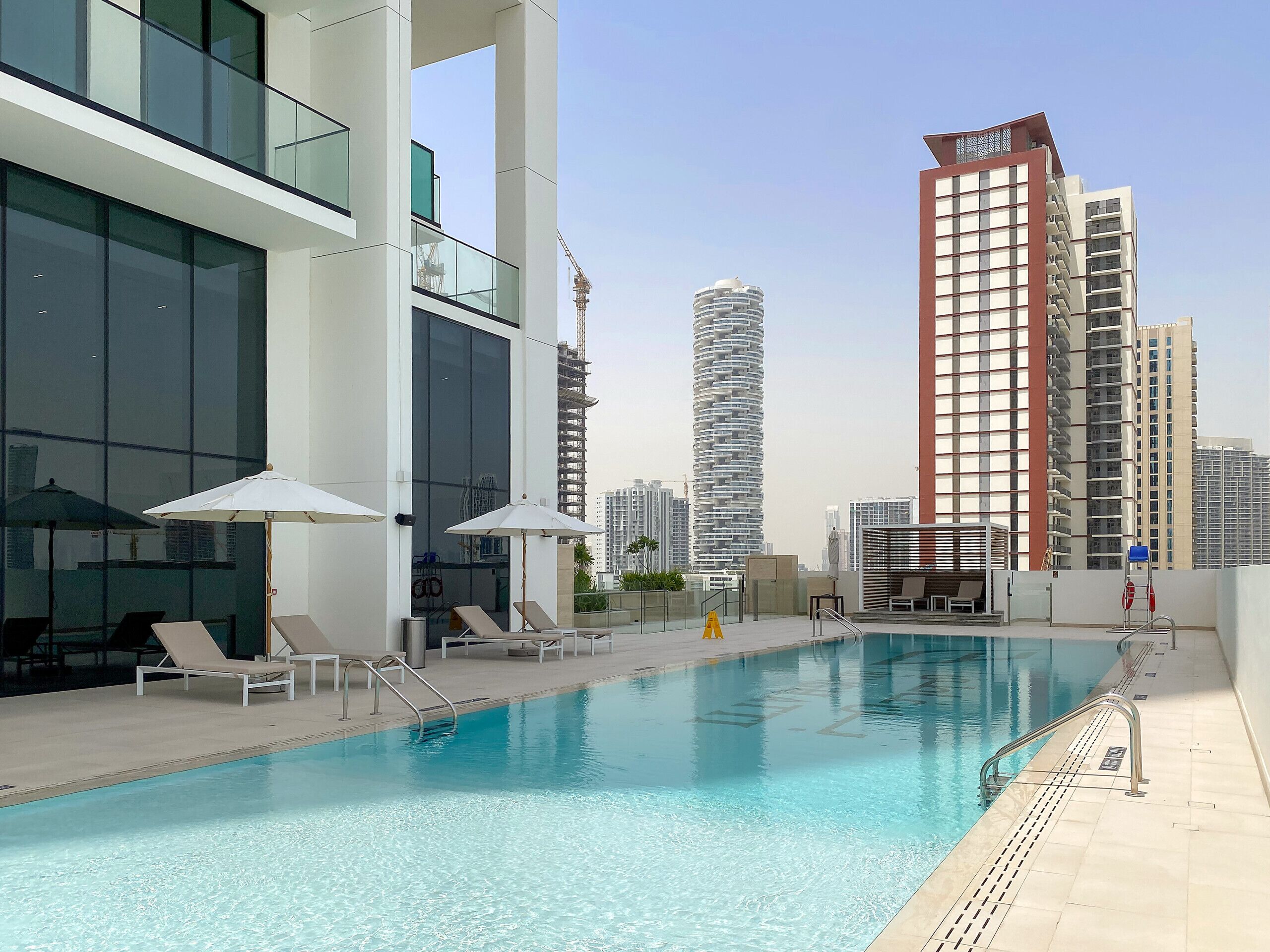 Una piscina sul tetto con poltrone a sdraio e ombrelloni, circondata da edifici moderni e un cielo sereno.