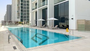 A modern hotel pool area with a clear blue pool, sun loungers, and umbrellas. - Cozy Stylish 1 BD in JVC (Dubai)