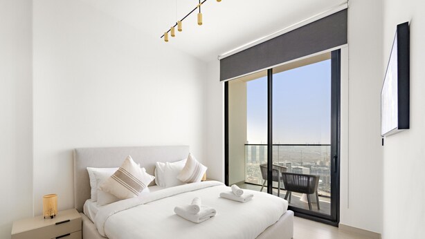 A modern hotel room with a large bed, a nightstand, a lamp, and a balcony with a city view. - Cozy Stylish 1 BD in JVC (Dubai)