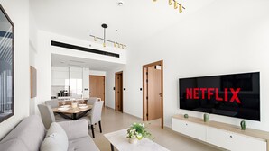 A modern living room with a large flat-screen TV, a sofa, a dining table, and a kitchen area in the background. - Cozy Stylish 1 BD in JVC (Dubai)