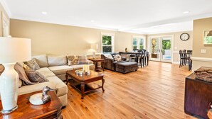 A spacious living room with a wooden floor, a sofa set, a dining table, and a large window providing natural light.