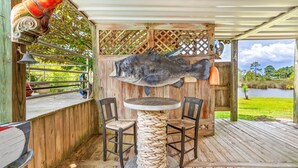 An outdoor wooden deck with a round table and two chairs, a large fish sculpture, and a bell.