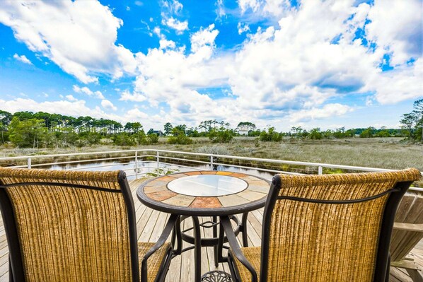 A deck with wicker chairs and a glass table, overlooking a natural landscape with trees and a body of water.