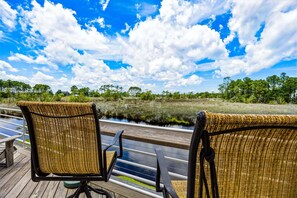 Two wicker chairs on a wooden deck overlooking a water body with a view of a forested area under a partly cloudy sky.