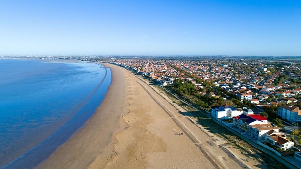 An aerial view of a coastal town with a long sandy beach, residential buildings, and a clear blue sky.