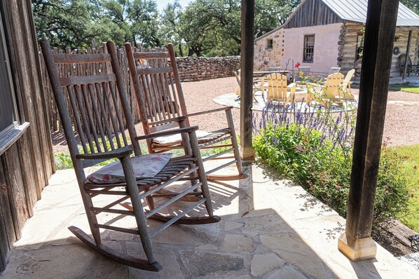 A rustic wooden rocking chair with a cushion, positioned on a stone patio with a view of a stone building and a garden with purple flowers.