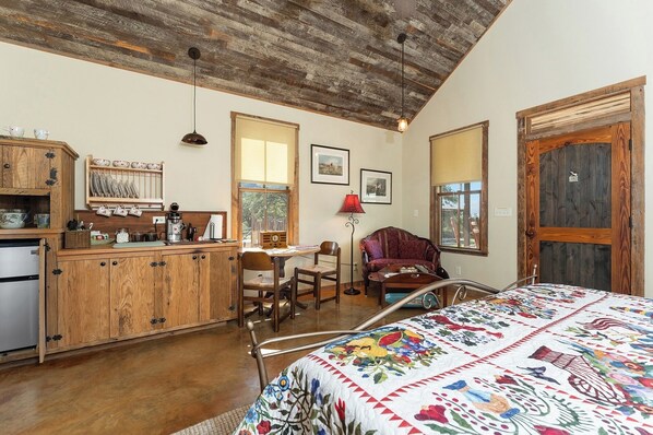 A rustic room with a wooden ceiling, a bed with a colorful quilt, a dining area with a wooden table, and a kitchen with wooden cabinets and a coffee machine. - Miss Thelma's | Cozy Country Retreat (Fredericksburg)