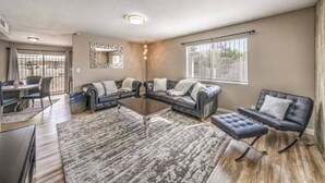 Un salón espacioso con un sofá modular grande, una mesa de centro y una televisión. Hay una zona de comedor con una mesa y sillas, y una ventana con persianas.