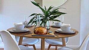 Une table de salle à manger ronde, entourée de chaises blanches, avec une miche de pain, des bols blancs et des verres à vin.