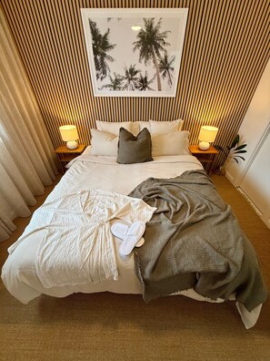 Ett hotellrum med en säng, två sänglampor, en inramad tavla med palmer och ett fönster med gardiner.