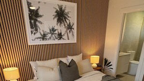 Une chambre d’hôtel avec un lit, deux tables de chevet avec des lampes, un grand tableau encadré au mur et une salle de bain visible à travers une porte ouverte.
