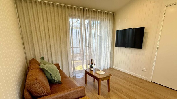 Un salon avec un canapé en cuir marron, une table basse en bois, un petit bol en bois, une bouteille de champagne et un téléviseur fixé au mur.