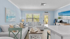 Un salon avec un mur bleu clair, un canapé agrémenté de coussins à motifs d’étoiles, une table basse sur laquelle sont posées des serviettes roulées, et un tableau représentant une scène de plage.