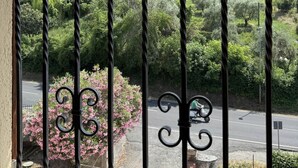 Un balcon doté d’une balustrade en fer forgé, donnant sur une route et offrant une vue sur une colline parsemée d’arbres, avec un bâtiment visible au loin.