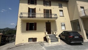 Un immeuble de deux étages doté d’un balcon, une voiture garée devant, et un ciel dégagé.
