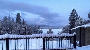 Une clôture en bois recouverte de neige, avec en arrière-plan un lac et des arbres.