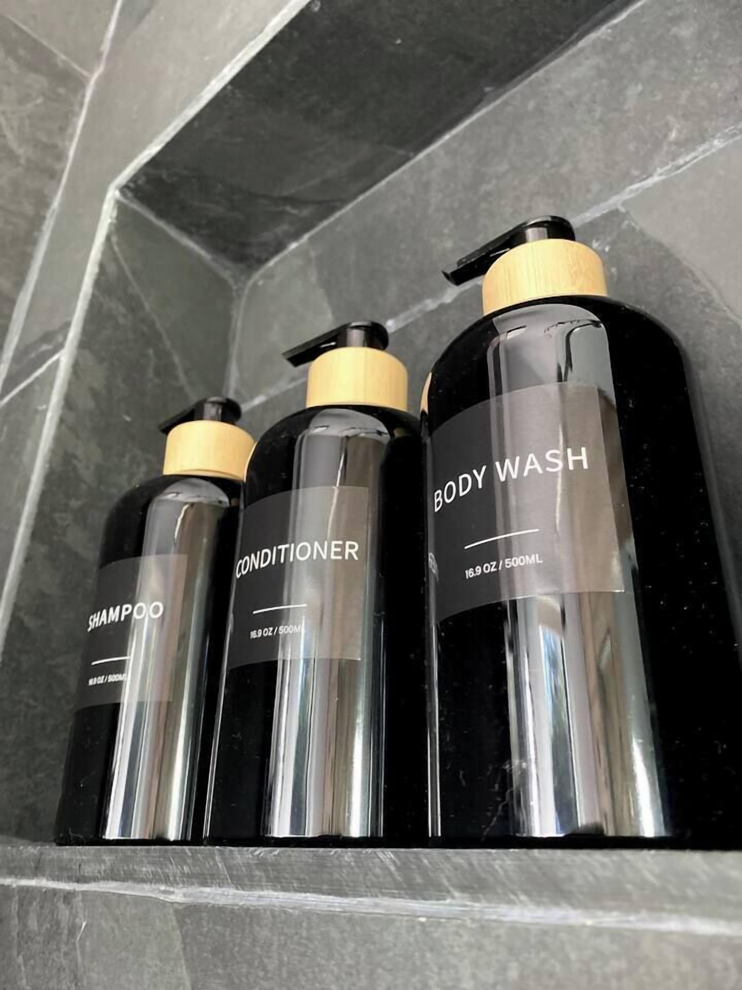 Trois bouteilles noires avec des bouchons dorés, étiquetées « GEL DOUCHE » et « APRÈS-SHAMPOING », sont posées sur une étagère en marbre.