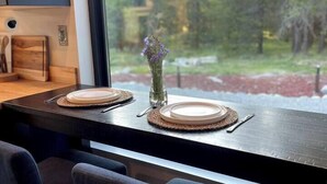 Un coin repas avec une table dressée pour deux personnes, un vase rempli de fleurs et une vue sur les arbres par la fenêtre.