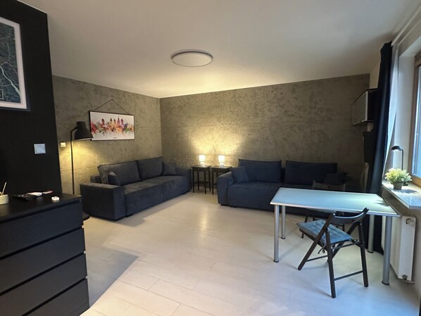 A modern living room with a grey sofa, a small table, and a chair. There is a painting on the wall and a lamp on a side table.