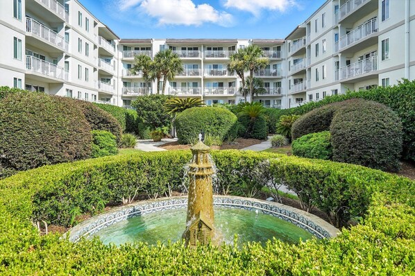 A multi-story hotel with a central courtyard featuring a fountain, manicured hedges, and palm trees.