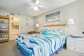 A bedroom with a bed, a ceiling fan, a nightstand, a lamp, a bookshelf, and a painting on the wall.