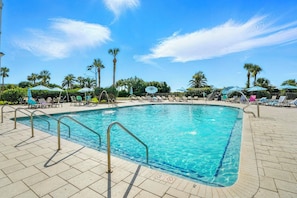 A rectangular swimming pool with clear blue water, surrounded by lounge chairs and umbrellas, with palm trees and a clear sky in the background.