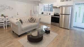 A modern kitchen with a stainless steel refrigerator, a dining area with a white table and chairs, and a living room with a beige sofa and glass coffee table. - Kam Residence  (Chilliwack)