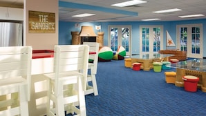 Zona de juegos infantiles con sillas blancas, pufs de colores y una casita.