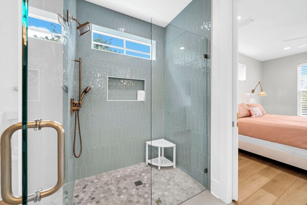 A modern bathroom with a glass shower enclosure, a small white shelf, and a view of a bedroom with a bed and a nightstand.
