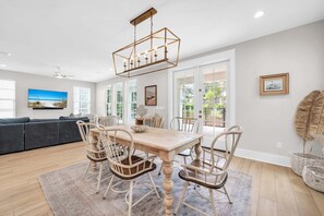 A dining area with a wooden table and chairs, a ceiling light fixture, and a view through large windows.