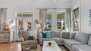 Un salon avec un fauteuil beige, un canapé gris et une table basse blanche sur laquelle on peut voir des fruits. On y trouve également des rideaux, une lampe et une vue sur l’extérieur.