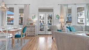 Un vaste salon doté d’un plancher en bois, un coin repas avec une table et des chaises blanches, un canapé avec des coussins bleus et un lustre.