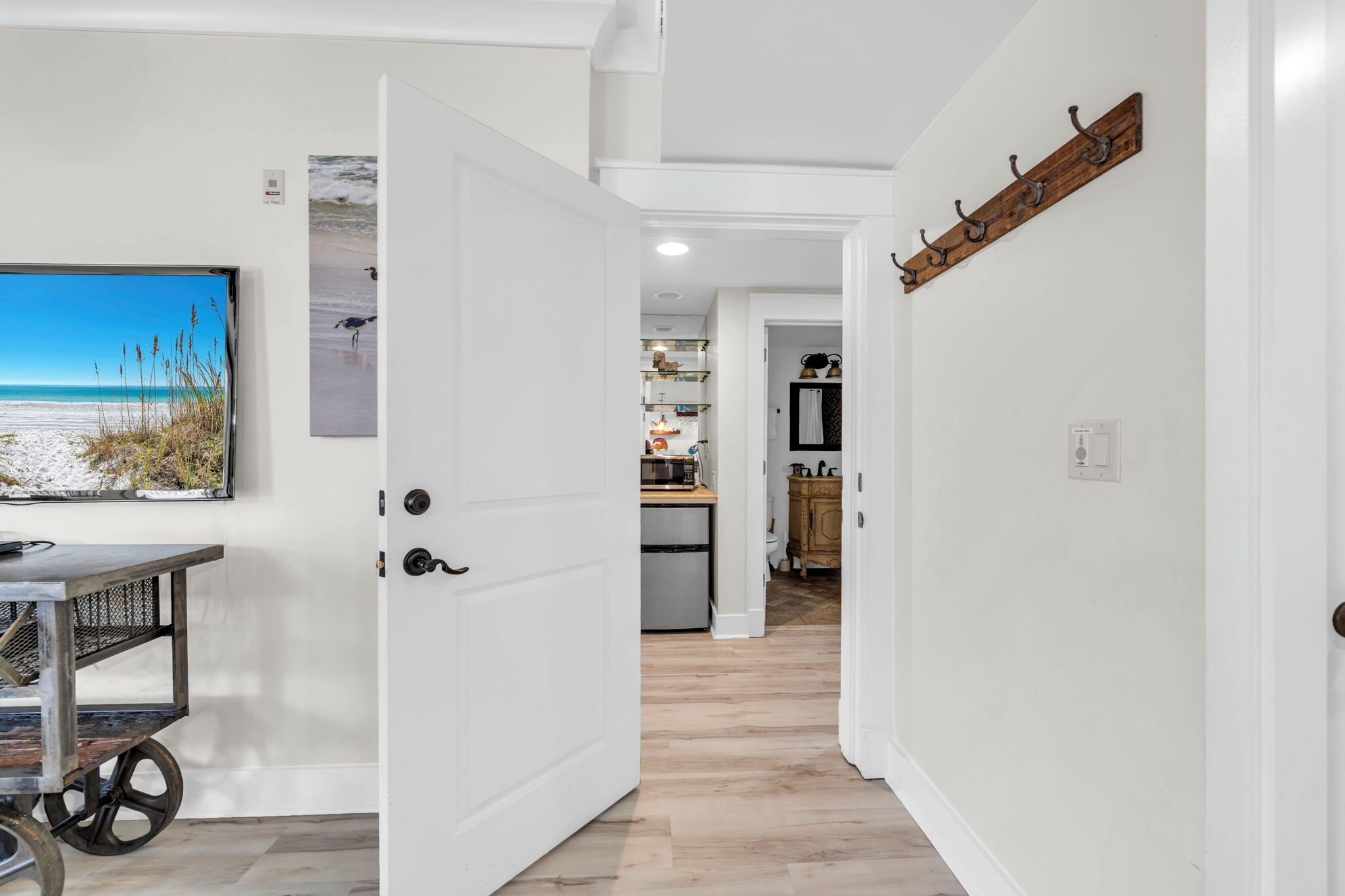 A hallway leading to a kitchen with a wooden floor, a mounted TV, and a wall-mounted coat rack.