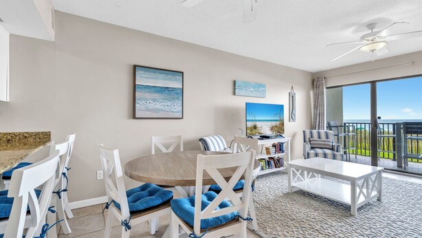 A spacious living area with a round table, white chairs, and a ceiling fan. There is a large window offering a view of the ocean. - Emerald Towers West 1005- Beach Front with Gulf View Balcony! (Fort Walton Beach)