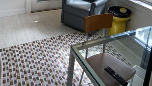 A room with a glass door, a blue armchair, a yellow stool, a glass table with a remote control, and a patterned rug.