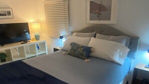 A bedroom with a bed, a TV on a stand, a bedside table with a lamp, and a window with blinds.