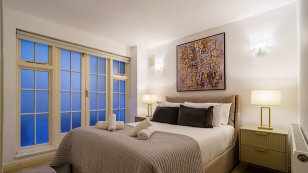 A neatly arranged bedroom with a large bed, two bedside tables with lamps, a window with blue-tinted glass, and a wall-mounted abstract painting.