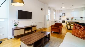 A modern living room with a wooden coffee table, a brown armchair, and a sofa. A flat-screen TV is mounted on the wall, and there is a kitchen area visible in the background.