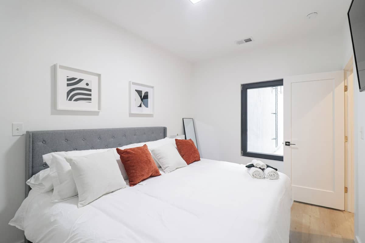 A modern bedroom with a grey upholstered headboard, white bedding, and two abstract wall art pieces.