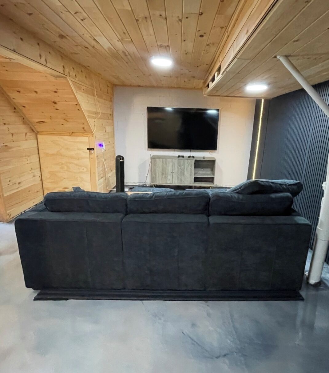 A modern living room with a dark grey sectional sofa, a flat-screen TV mounted on the wall, and wooden paneling on the ceiling.